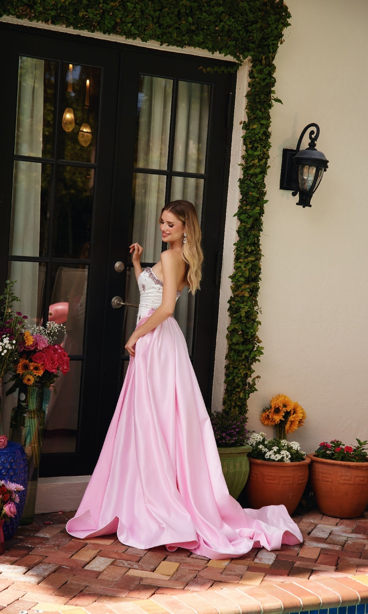 A woman in the Ava Presley 40088 long strapless prom dress with a train stands by a glass door, surrounded by potted flowers and lush greenery on a brick patio.